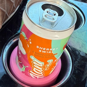 Könnte beinhalten: Eine Getränkedose "Sherbet Swirl" in einem Getränkehalter im Auto. Die Dose hat ein farbenfrohes Etikett mit orangefarbenen, rosa und weißen Grafiken und Text. Sie steht in einem rosa Schaumstoff-Einsatz in einem silbernen Getränkehalter.