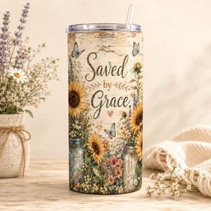 May include: A cylindrical tumbler with a clear lid and straw, featuring a rustic wood-grain background, sunflowers, wildflowers, and butterflies. The text "Saved by Grace" is printed in a dark script font with a small pink heart below it.