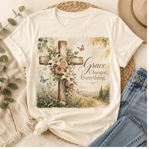 May include: A cream-colored short-sleeve t-shirt featuring a graphic print of a wooden cross adorned with pink roses, white lilies, and green foliage. Butterflies are depicted around the cross, with the text Grace Changes Everything.