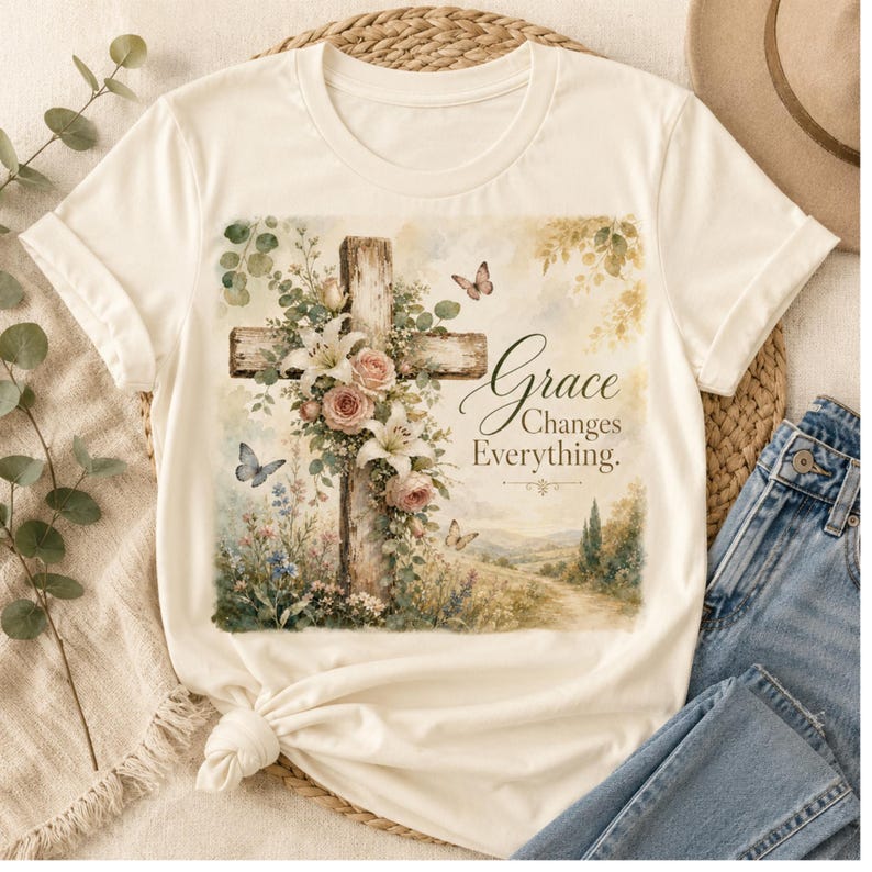 May include: A cream-colored short-sleeve t-shirt featuring a graphic print of a wooden cross adorned with pink roses, white lilies, and green foliage. Butterflies are depicted around the cross, with the text Grace Changes Everything.