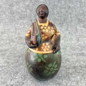 May include: A ceramic figurine of a person with dark skin, wearing a yellow top with white polka dots and a dark green skirt featuring light green floral patterns. The figure holds a small, smooth, dark-toned vessel in the right hand.