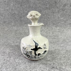 May include: A cream-colored ceramic decanter featuring a textured surface and a sculpted stopper shaped like a stylized face. The body of the vessel is decorated with a dark, hand-painted graphic depicting a bird in flight over a landscape.