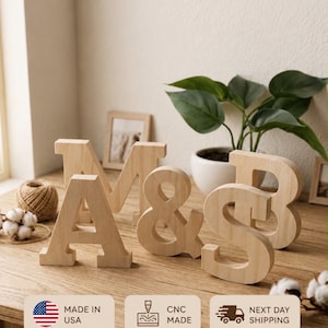 Op de afbeelding: Vrijstaande houten letters A, M, &, en B met een lichte natuurlijke houtnerfafwerking. De letters zijn in een groep op een houten oppervlak geplaatst. De afbeelding bevat tekstoverlays met Made in USA, CNC Made, Next Day Shipping, Natural Wood, Sustainably Sourced, Perfect for Any Space en Great Gift Idea.