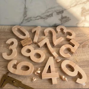 Puede incluir: Una colección de diez números de madera de color claro del cero al nueve, con superficies lisas y bordes redondeados. Se incluye un pequeño bloque de madera rectangular con el texto HAND CRAFTED, junto con una herramienta de calibre de metal.