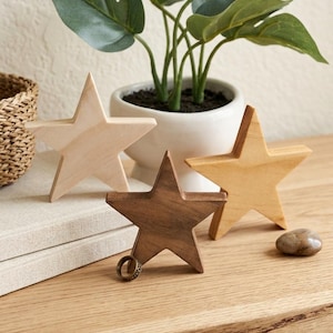May include: Four wooden star decorations in varying shades of brown and tan are arranged on a wooden surface. A small potted plant with green leaves sits in the background. A ring rests on one of the stars.