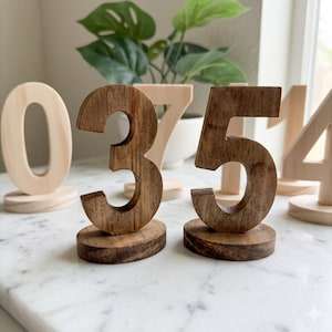 May include: A collection of freestanding wooden numbers, including a dark brown stained number three and number five in the foreground, with lighter natural wood numbers zero, seven, one, and four positioned in the background on a white surface.