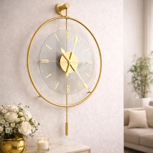 May include: A gold-colored, modern wall clock with a clear glass face. The clock features gold hands and a pendulum. The clock is mounted on a white wall. The clock's diameter is approximately 18 inches.