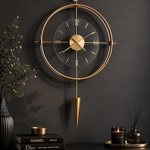May include: A gold-coloured, modern wall clock with a circular frame and a clear glass face. The clock features gold hands and hour markers, with a conical pendulum. The clock is mounted on a dark grey wall, with decorative items on a black surface below.