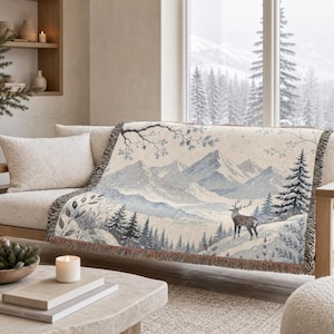 May include: A woven throw blanket featuring a detailed winter landscape scene with snow-capped mountains, evergreen trees, and a standing deer. The design is rendered in shades of cream, light blue, and dark grey with a fringed edge.