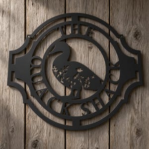 Pode incluir: Uma placa de parede circular em metal preto com a silhueta de um pato que apresenta recortes florais no corpo. O texto THE QUACK SHACK está integrado na moldura circular que rodeia o pato.