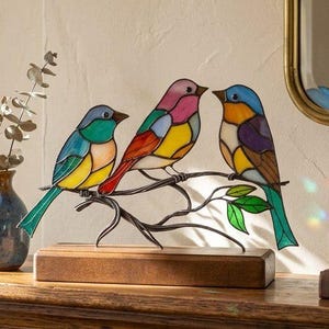 May include: A stained glass sculpture of three colorful birds perched on a branch. The birds feature vibrant hues of blue, yellow, pink, and green. The sculpture sits on a wooden base, adding a touch of nature-inspired decor.