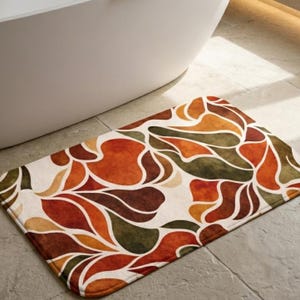 May include: A rectangular bath mat with a floral pattern in shades of orange, brown, and green. The mat is placed on a grey tiled floor near a white bathtub. The design features abstract leaf-like shapes.