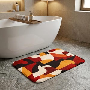 May include: A rectangular bath mat featuring an abstract geometric pattern in red, orange, black, and cream. The mat is positioned on a grey tiled floor in front of a white bathtub, providing a vibrant accent to the bathroom.