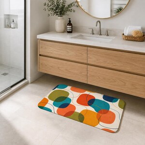 May include: A rectangular bath mat featuring a cream-colored background with an overlapping pattern of rounded shapes in shades of orange, teal, olive green, and dark blue, accented by thin, dark, continuous curved lines throughout the design.