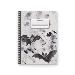 Op de afbeelding: Spiraalgebonden notitieboek met een grijswaardenontwerp met vleermuizen, bloemen en spinnenwebben. De omslag bevat het woord "NOTEBOOK" in een rechthoekig label. Het ontwerp van het notitieboek heeft een vintage, gotische esthetiek.