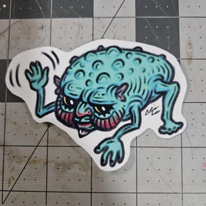 May include: A sticker depicting a teal cartoon creature with a bumpy head, small limbs, and a red and pink face. The creature is waving with one hand and has yellow eyes. The sticker is on a grey and white grid.