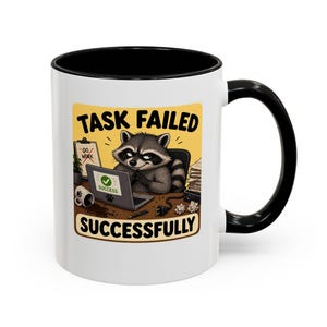 May include: White ceramic mug with a black interior and handle. The mug features a cartoon raccoon working on a laptop with the text "TASK FAILED SUCCESSFULLY". The design includes a yellow background and office-themed elements.