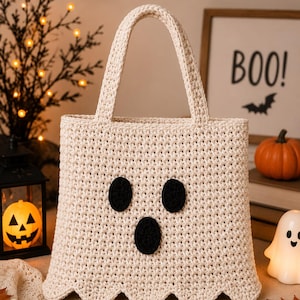 May include: A cream-colored crocheted tote bag featuring a ghost face design with two black oval eyes and one black oval mouth. The bag has a scalloped bottom edge and two matching cream-colored handles, crafted with a textured stitch pattern.