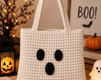 Puede incluir: Un bolso de mano tejido a ganchillo en color crema con un diseño de cara de fantasma que incluye dos ojos ovalados negros y una boca ovalada negra. El bolso tiene un borde inferior festoneado y dos asas a juego en color crema.