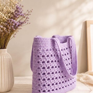 May include: A light purple crochet tote bag featuring a repeating openwork square pattern throughout the body and two matching solid crochet handles. The bag is constructed with a dense, textured stitch at the base and top rim for structure.
