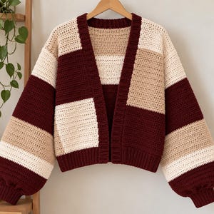 May include: A long-sleeved, open-front crochet cardigan featuring a color-blocked pattern in deep burgundy, cream, and beige. The garment has a ribbed collar and cuffs, with a loose, boxy fit and dropped shoulders, constructed from yarn.