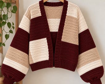 Puede incluir: Un cárdigan de ganchillo de manga larga y frente abierto con un patrón de bloques de color en burdeos intenso, crema y beige. La prenda presenta cuello y puños acanalados, un corte holgado y hombros caídos, tejido en hilo.