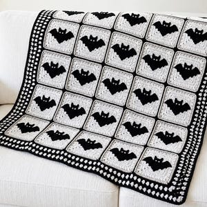 May include: A handmade crochet blanket featuring a repeating pattern of black bat motifs centered on light gray square panels. The blanket is bordered by a thick black crochet frame with a textured, repeating stitch pattern along the edges.