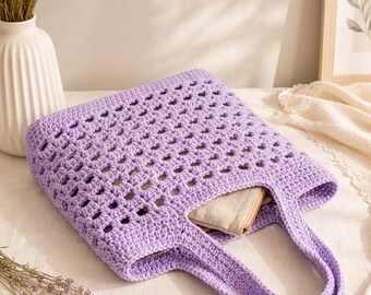 Puede incluir: Un bolso tote de ganchillo de color morado claro con un patrón de cuadrados calados en el cuerpo principal, una base sólida y dos asas integradas. Un pequeño objeto de tela beige está metido dentro de la parte superior abierta del bolso de hilo.