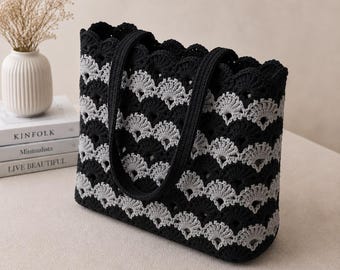 Puede incluir: Un bolso tote rectangular hecho de hilo de ganchillo con un patrón repetitivo de conchas festoneadas en franjas horizontales alternas de negro y gris claro. El bolso incluye dos asas largas de ganchillo negro unidas al borde superior.