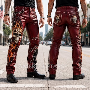 Puede incluir: Pantalones de cuero rojo oscuro con tachuelas metálicas plateadas a lo largo de las costuras, bolsillos con cremallera y un estampado gráfico de una calavera rodeada de llamas naranjas y amarillas en el frente y los bolsillos traseros. Texto AMERICASTARS.