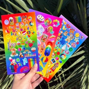 May include: A hand holds three sheets of colorful stickers with a rainbow gradient background. The stickers feature various designs, including rainbows, stars, cupcakes, and teddy bears. One sheet has the text "be kind".