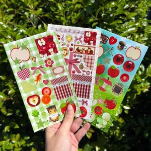 May include: Three sheets of decorative stickers featuring apple and fruit designs. The stickers include red apples, bows, and gingham patterns in green and red. Other designs include a goldfish, cat, and coffee cup.