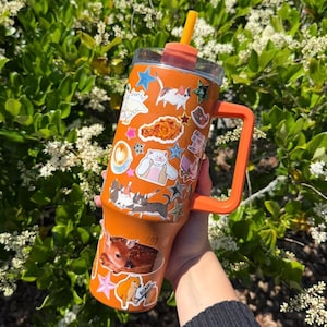 May include: An orange insulated tumbler with a handle and a straw, featuring a variety of stickers including cats, a deer, a fried chicken piece, a coffee cup, and stars. The tumbler has a clear lid and a bright orange straw extending from the top.