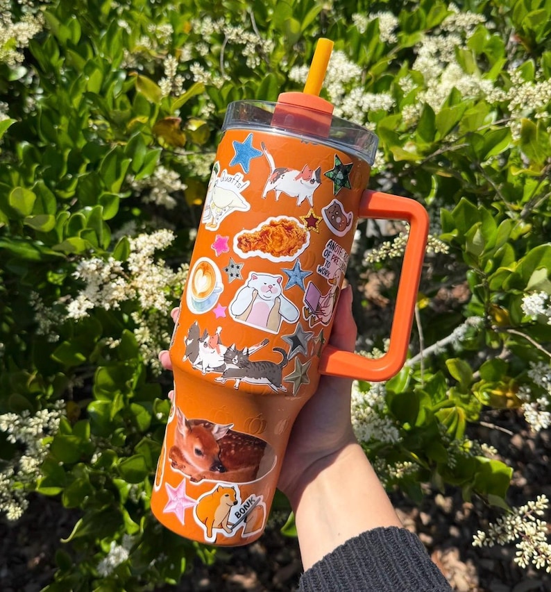 May include: An orange insulated tumbler with a handle and a straw, featuring a variety of stickers including cats, a deer, a fried chicken piece, a coffee cup, and stars. The tumbler has a clear lid and a bright orange straw extending from the top.