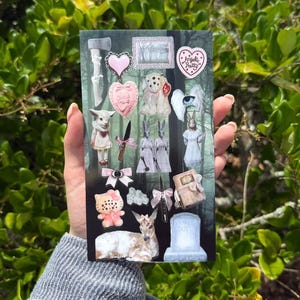 May include: A rectangular sticker sheet featuring various illustrations including a grey axe, pink hearts, a white creature with multiple eyes, a knife with a pink bow, twin rabbits, a winged eye, a cat figure, a deer, and a tombstone labeled DOLL.
