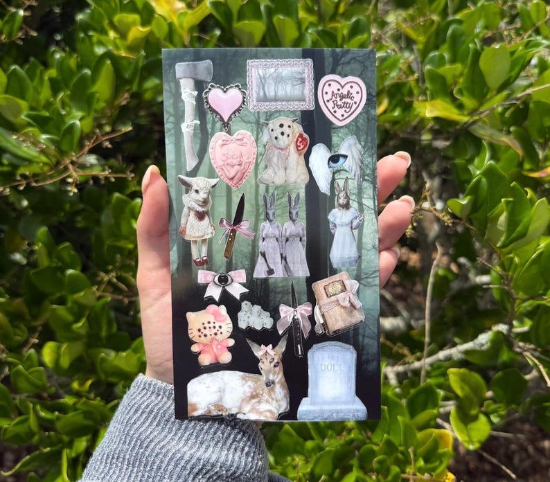 May include: A rectangular sticker sheet featuring various illustrations including a grey axe, pink hearts, a white creature with multiple eyes, a knife with a pink bow, twin rabbits, a winged eye, a cat figure, a deer, and a tombstone labeled DOLL.