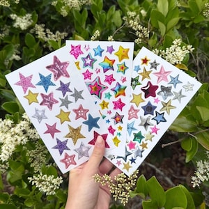 May include: Three sheets of star-shaped stickers in various colors and textures, including glitter and metallic finishes. The stickers are in shades of pink, blue, yellow, and silver, with some featuring rainbow gradients. The sheets are held against a backdrop of green foliage.