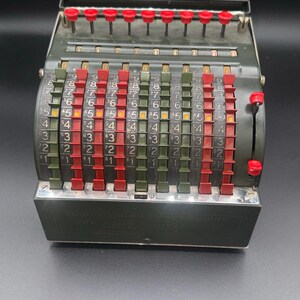 Pode incluir: Uma antiga máquina de somar Todd Visible com caixa de metal verde-escura. O dispositivo possui várias colunas verticais de alavancas deslizantes em vermelho e verde-oliva, numeradas de um a nove, com uma fileira de botões vermelhos no topo.