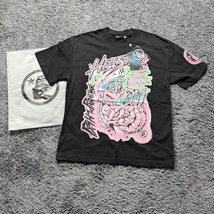 May include: A black short-sleeved t-shirt featuring a large graphic print on the front with the text "HELLSTAR" and "NO GUTS NO GLORY!" alongside a pink brain illustration. A matching white plastic bag with a circular logo is positioned to the left.