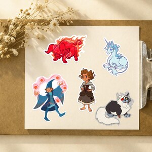Puede incluir: Un conjunto de pegatinas de tem&aacute;tica fant&aacute;stica. Incluyen un toro rojo en llamas, un unicornio azul, un mago, una ni&ntilde;a con un vestido y un gato con pelaje blanco y negro. Las pegatinas est&aacute;n sobre papel blanco, sujetas por una pinza.