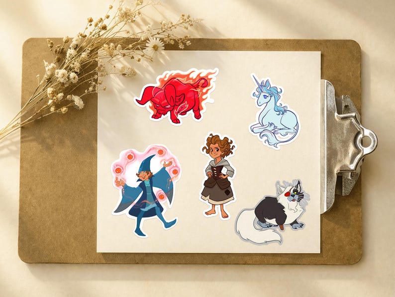 Puede incluir: Un conjunto de pegatinas de tem&aacute;tica fant&aacute;stica. Incluyen un toro rojo en llamas, un unicornio azul, un mago, una ni&ntilde;a con un vestido y un gato con pelaje blanco y negro. Las pegatinas est&aacute;n sobre papel blanco, sujetas por una pinza.