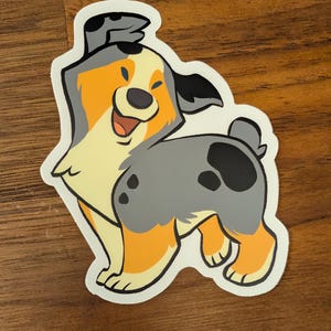 May include: A die-cut vinyl sticker featuring a stylized illustration of a blue merle Australian Shepherd dog. The dog has a cream-colored chest, orange legs and face accents, and grey fur with black spots. It is shown standing and looking back.