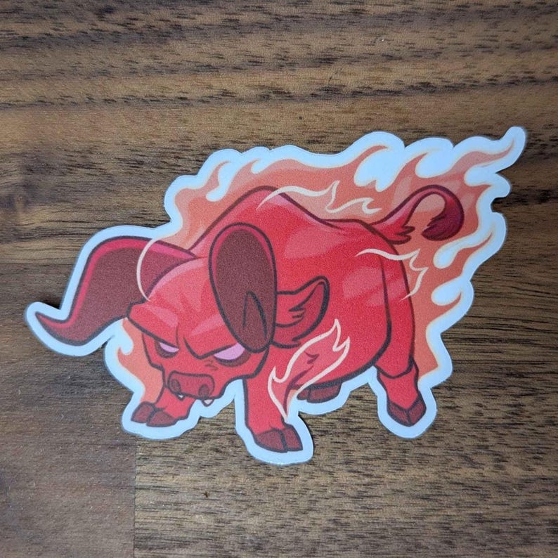 Puede incluir: Una pegatina con un toro rojo de dibujos animados con un aura de fuego. El toro tiene una expresi&oacute;n severa y est&aacute; rodeado de llamas naranjas y blancas. La pegatina est&aacute; sobre una superficie de madera.