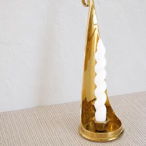 Puede incluir: Un candelabro de latón pulido con un panel trasero alto y curvado en forma de vela y una base circular. Una vela cónica blanca, en espiral y retorcida, está colocada en el centro del candelabro, descansando en una pequeña copa redonda.
