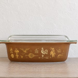 May include: A rectangular brown glass casserole dish with a clear glass lid. The exterior of the dish features a repeating gold-colored pattern of various kitchen and farm-themed icons, including a rooster, a coffee grinder, and a weather vane.