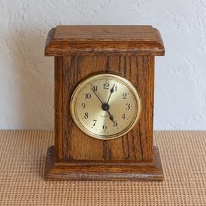 May include: A rectangular wooden mantel clock with a medium brown oak finish. The clock features a circular gold-toned face with black Arabic numerals, black hour and minute hands, and the text Quartz and Made in USA printed on it.