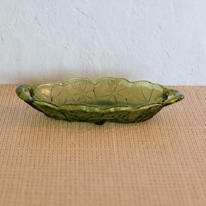May include: An oval-shaped olive green glass dish featuring a scalloped edge and integrated handles on both ends. The interior surface of the dish is decorated with a repeating embossed starburst pattern, and the base rests on small feet.