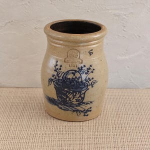 May include: A tan stoneware crock with a dark blue cobalt blue hand-painted design of a basket filled with berries. The vessel features a rounded rim, a stamped circular maker's mark, and the year 1991 printed in blue ink on the front surface.