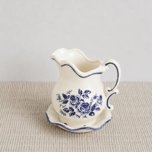 May include: A cream-colored ceramic creamer pitcher with a scalloped rim and a matching saucer base. The piece features a blue floral rose pattern on the front and a blue trim along the edges of the handle, rim, and base of the pitcher and saucer.