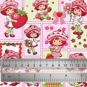 May include: A fabric print featuring Strawberry Shortcake characters in various poses with strawberries, flowers, and cupcakes, shown with a ruler measuring up to 5 inches and 13 cm to indicate the scale of the repeating pattern on the material.